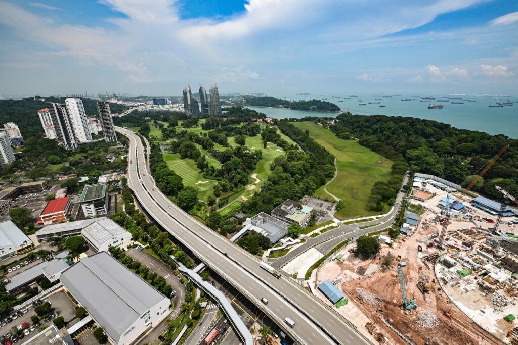 Aerial view of a coastal cityscape featuring a large green park or golf course surrounded by trees, a major highway curving through the scene, clusters of high-rise buildings, and a construction site. In the distance, numerous ships and boats are anchored in the ocean under a partly cloudy sky.